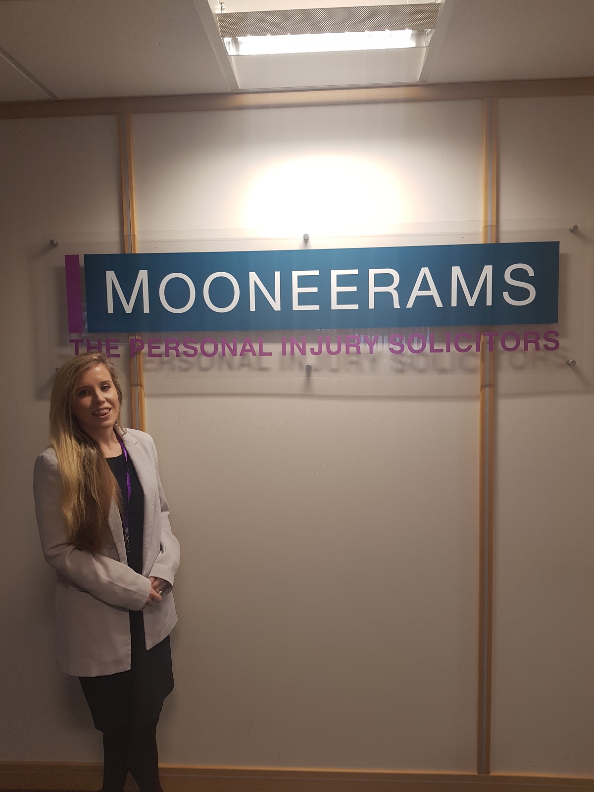 Female standing to the left of a sign with Mooneerams in white text on a blue background and ppersoanl injury solicitors written underneath the sign in pink lettering