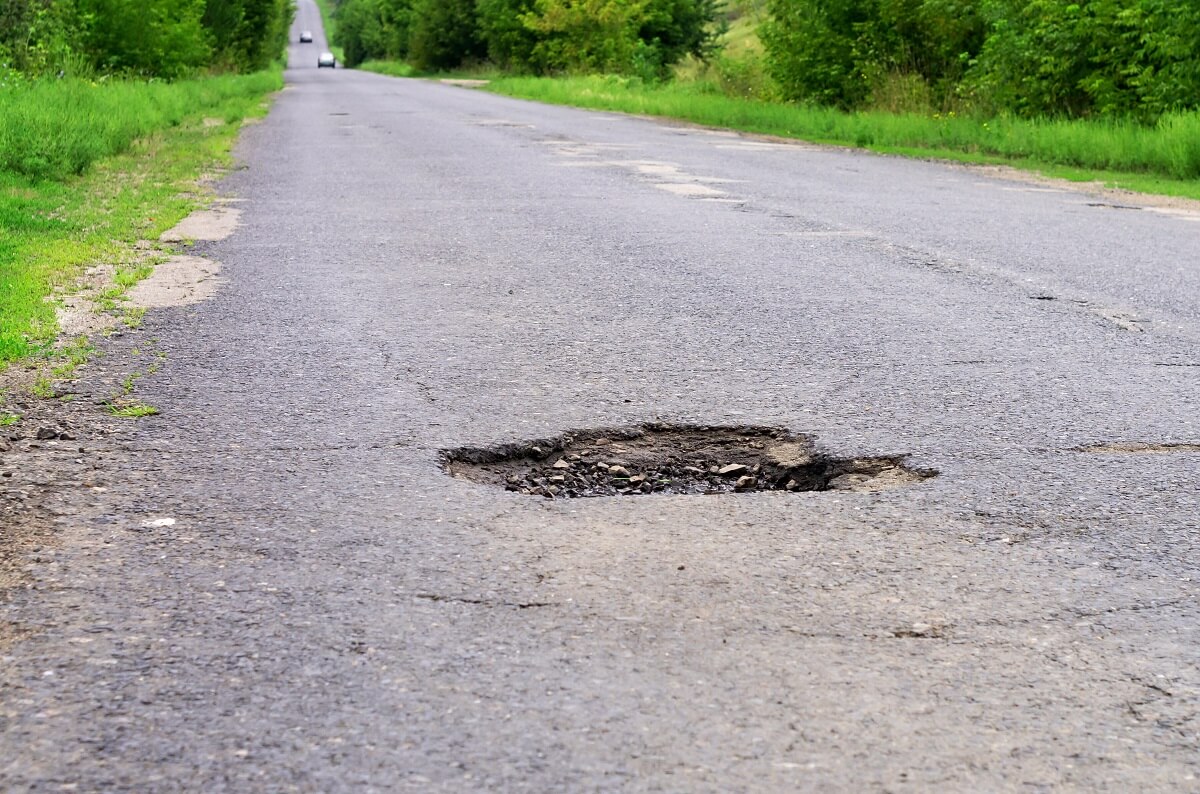 large hole in the middle of a grey road with green grass at either side of road