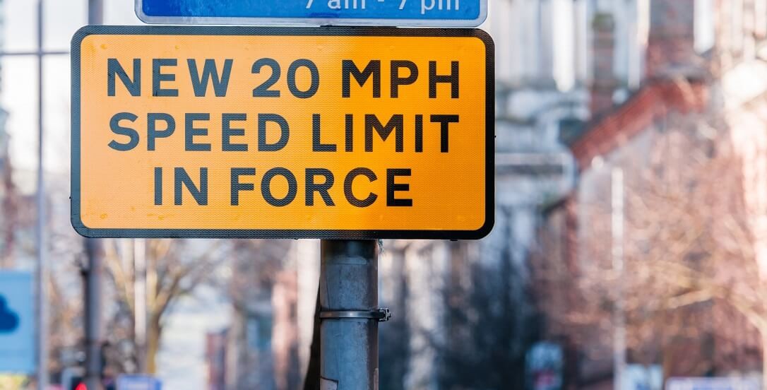 'New 20 mph speed limit in force' written in black on yellow street sign, with an urban street in the background